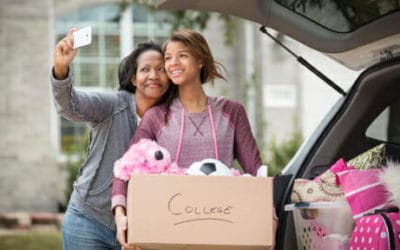 A mother and daughter take a selfie, the daughter holds a storage box marked college and there is a car behind with more boxes in the boot